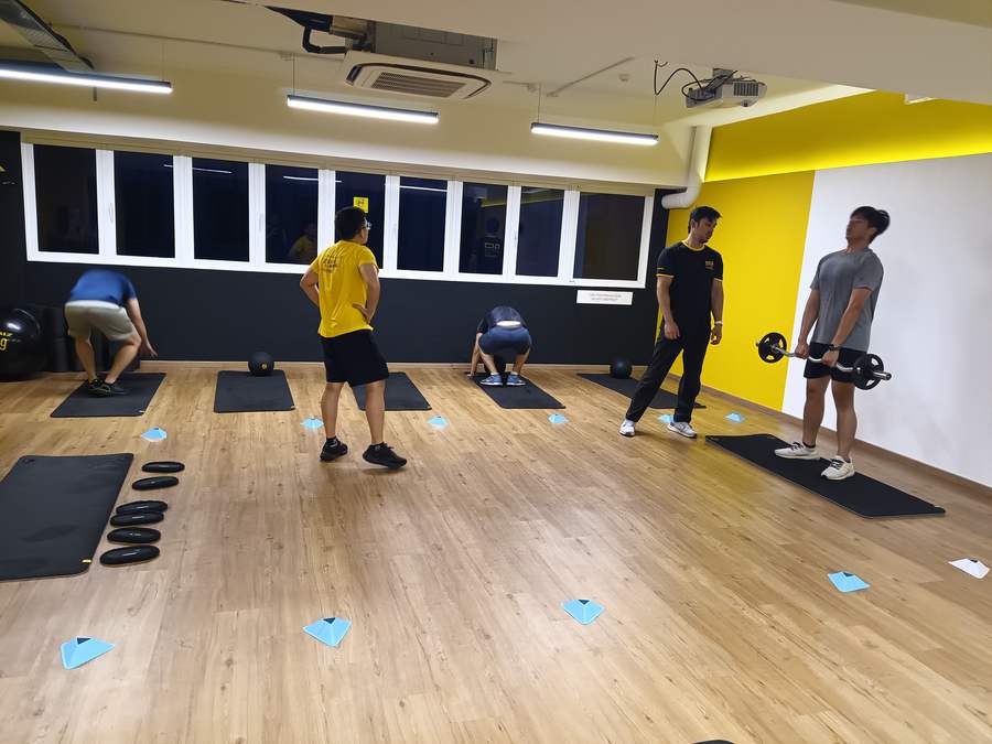 A coach leading a small evening class, members spread across mats with barbells and slam balls